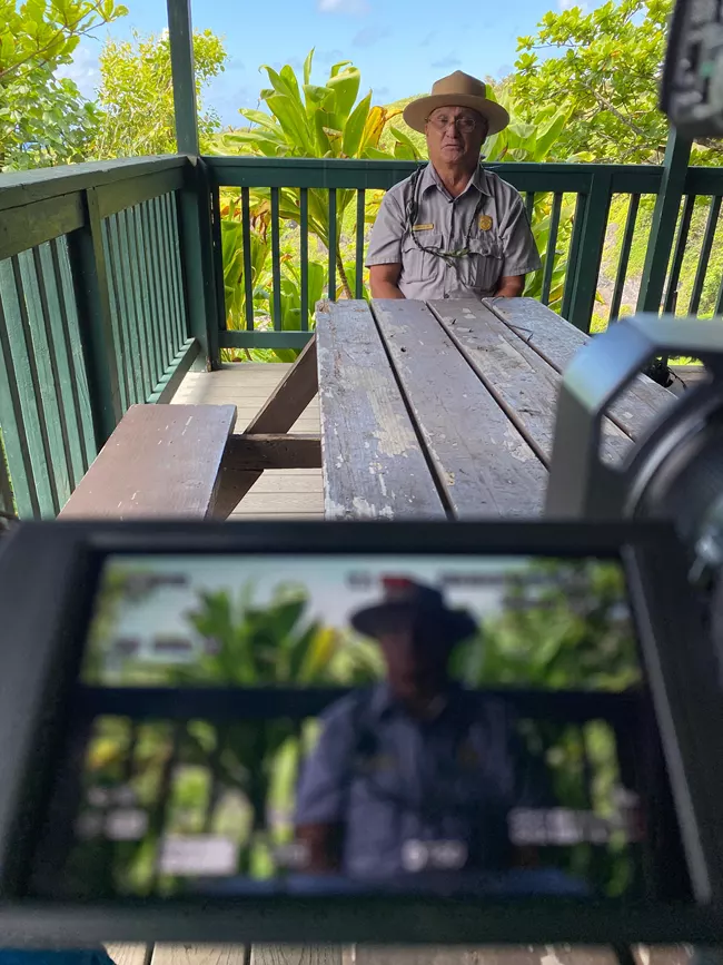 A man seated at a picnic table with a camera screen in the foreground that has a display showing him.