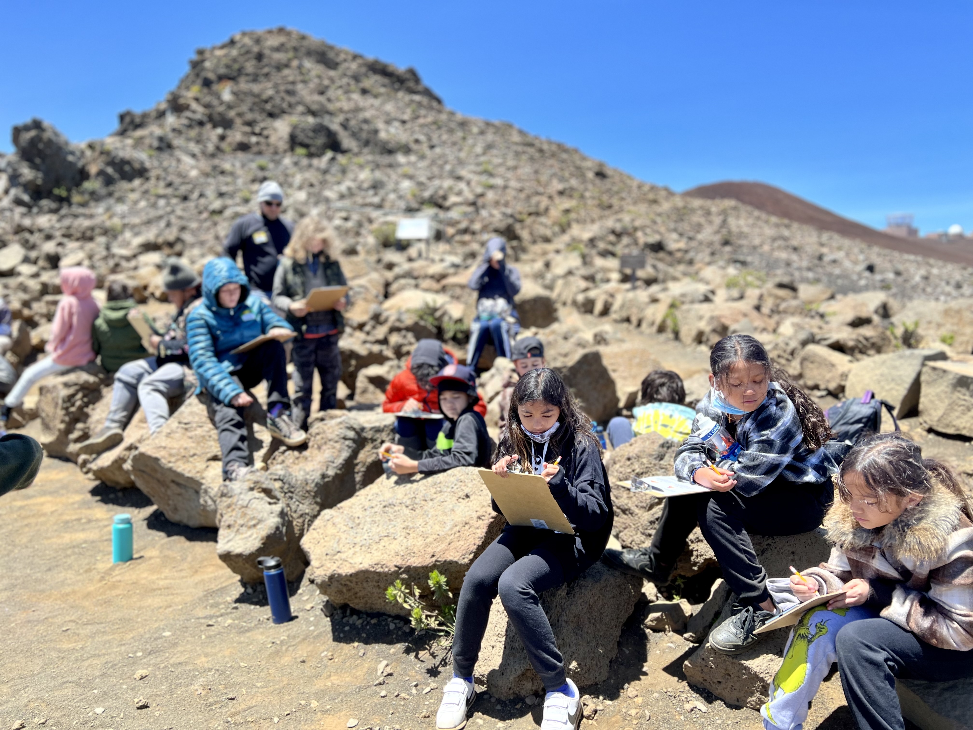 Elementary students sit on rocks outside working on clipboards