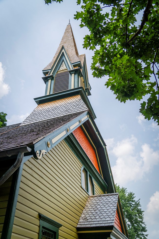 A tall church steeple, painted green, yellow, and orange, against a blue sky with a tree to the side.