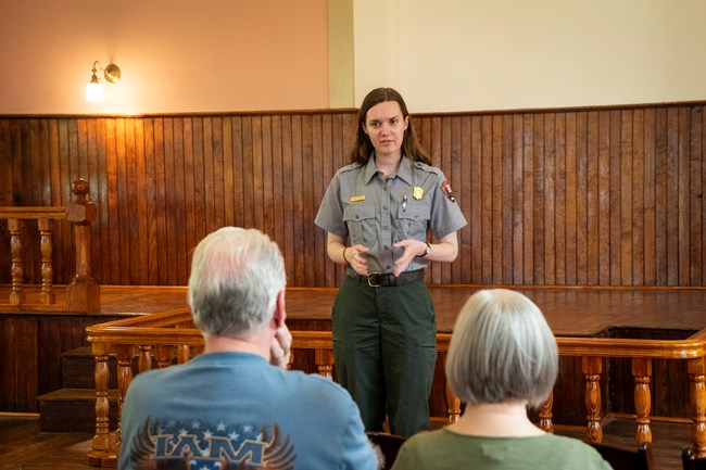 A woman in a ranger uniform stands inside a church and speaks to two seated visitors.