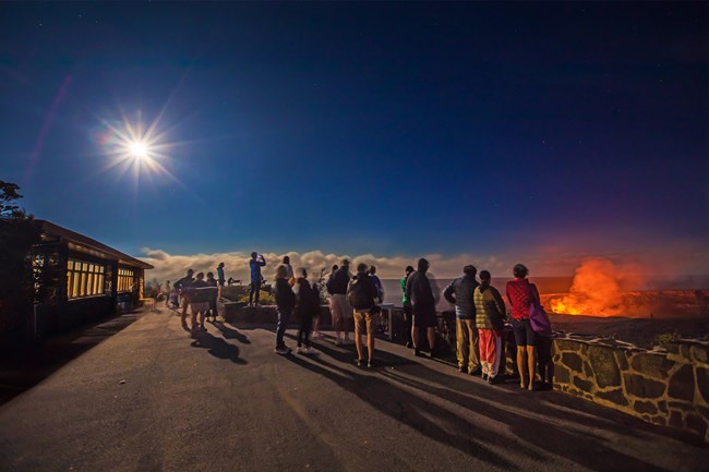 People standing at overlook looking at glowing lava in the distance with moon shining above.