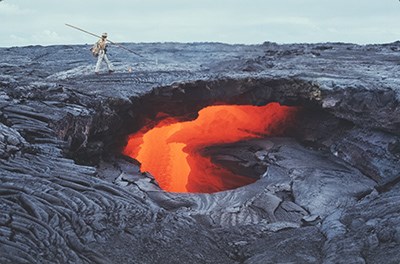 A hole in a solidified lava flow with glowing molten lava underneath, a man walks on top of the solidified flow