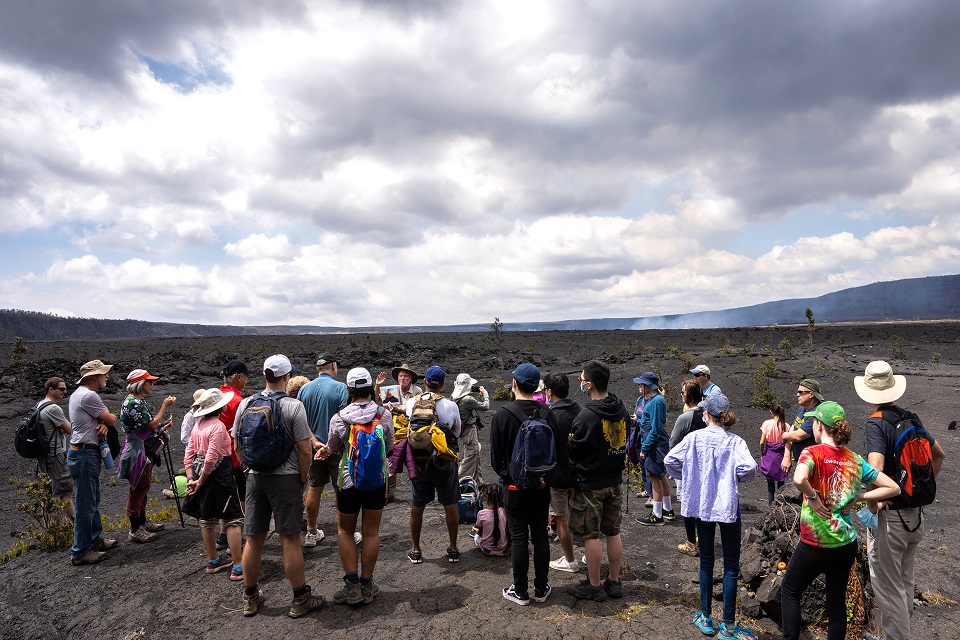 A volunteer park ranger holds a photo of an erupting volcano while standing on the caldera floor of Kilauea volcano with a few dozen visitors watching him