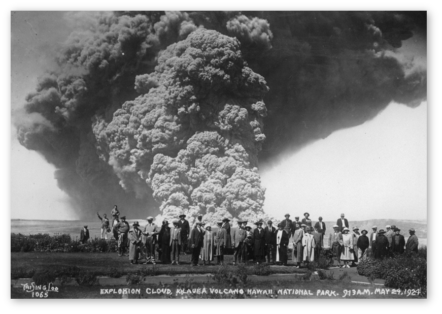 1924 Black and white photo of a crowd of people standing in front of an explosive volcano with a huge cloud of gas and ash billowing behind them upward