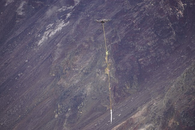 An unmanned aircraft system carrying a water container hanging by rope in a volcanic crater