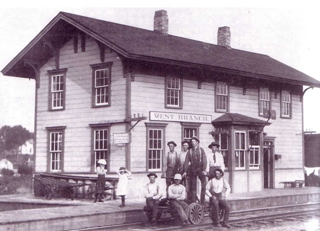 Men pose in front of a small town train station in the 1870s.