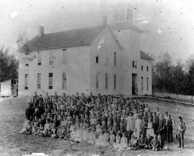 Several dozen schoolchildren of all ages in 1800s clothes pose for a photograph in front of their school building.