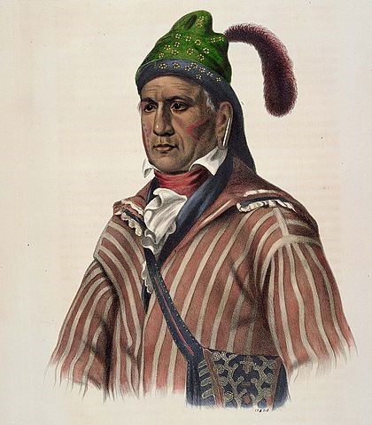portrait of Native American with red and white striped coat, satchel, green hat with feather, and face markings that appear to be tattoos on cheeks