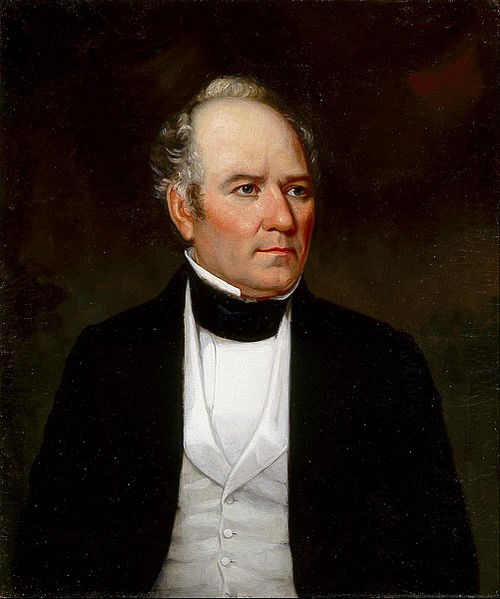 portrait of man from stomach up wearing black jacket, white vest and shirt, partially bald