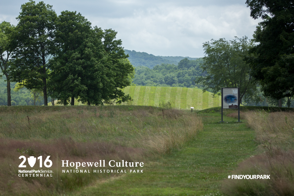 A grassy trail with a large sign and large mound in the view