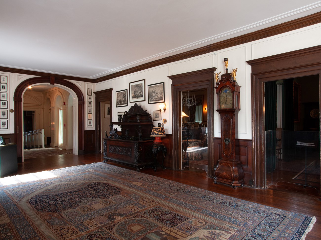 A room furnished with large oriental carpet, dark wood paneling and furniture, a grandfather clock, and many framed prints.