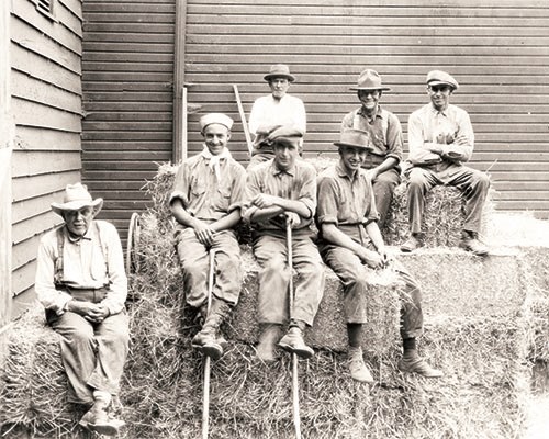 Seven men sitting on hay bales next to a barn.