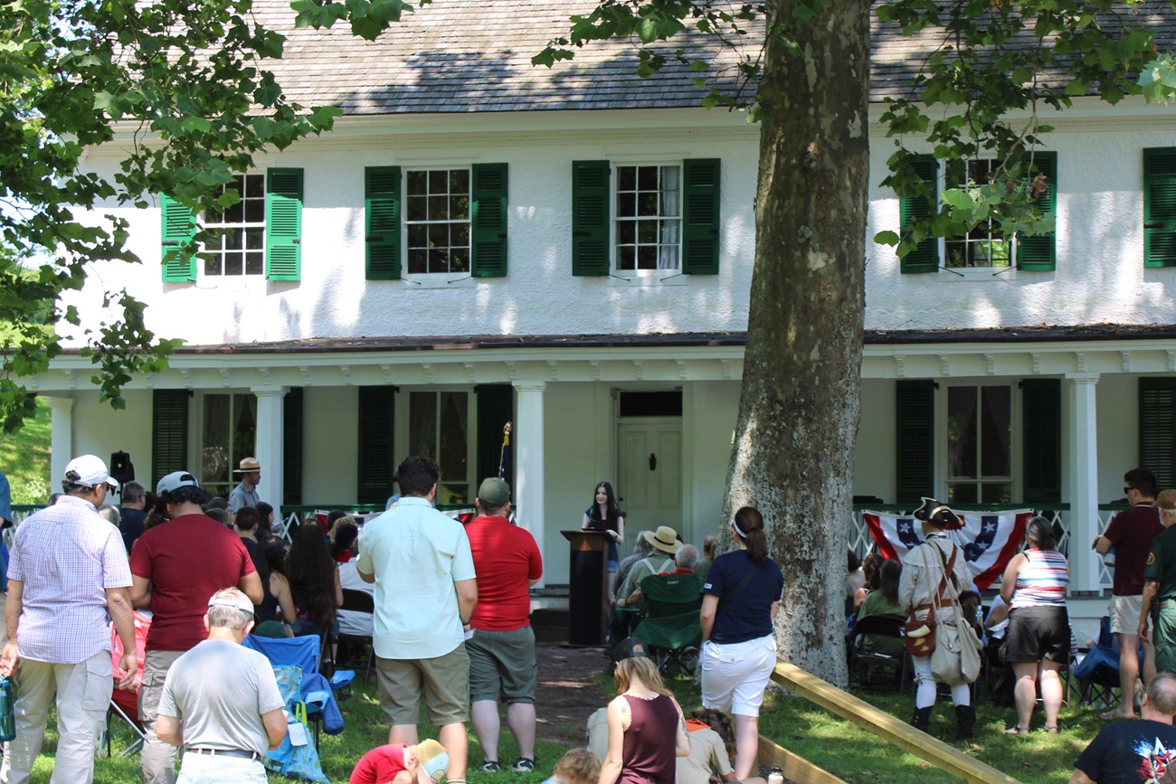 A group of people gather near the front of a large white building. A teenager speaks from the podium to the crowd.