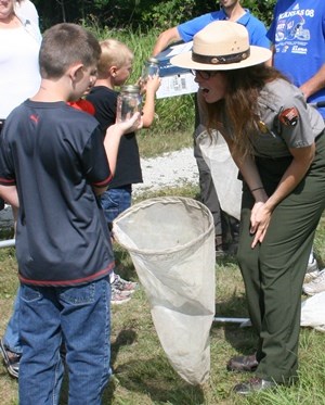 A boy holds a jar up for a female park ranger to inspect. He holds a butterfly net. There is a group in the background.