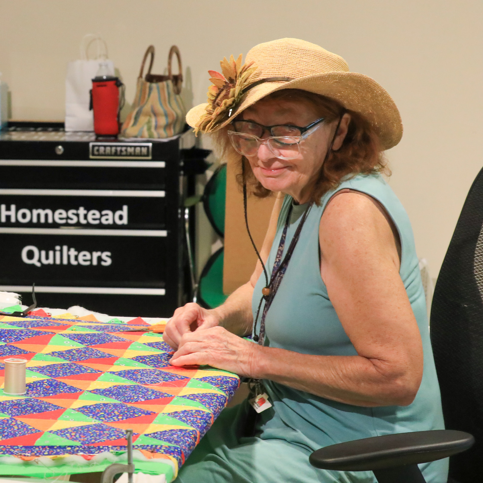 Woman with red hair under straw hat looks at camera through 2 sets of glasses while sewing.