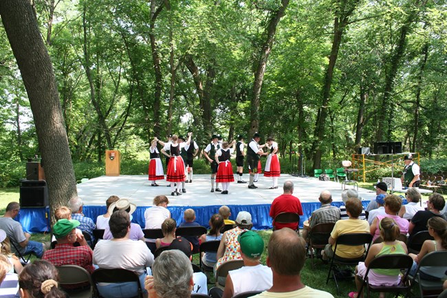 A group of dancers stands in a circle on an outdoor stage. The dancers wear traditional European dress. A crowd sits in the foreground and watches.