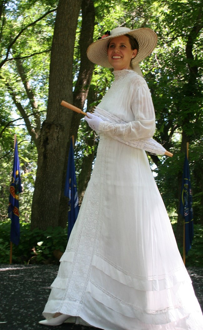 A woman in a long white dress and wide brimmed straw hat walks across a stage with a parasol under her arm.