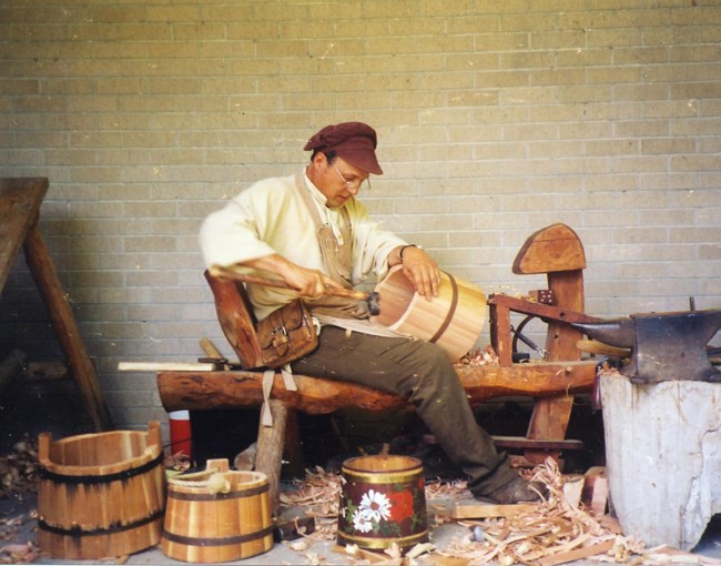 Man sitting on a bench holding a bucket on his lap with one hand and a tool in the other. Buckets are on the ground around him.