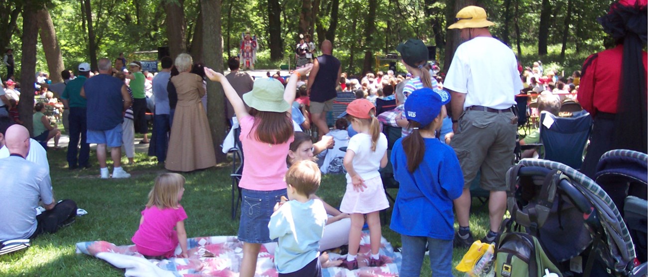 A crowd in the foreground watches a stage performance. A little girl in the foreground has both arms thrown in the air.