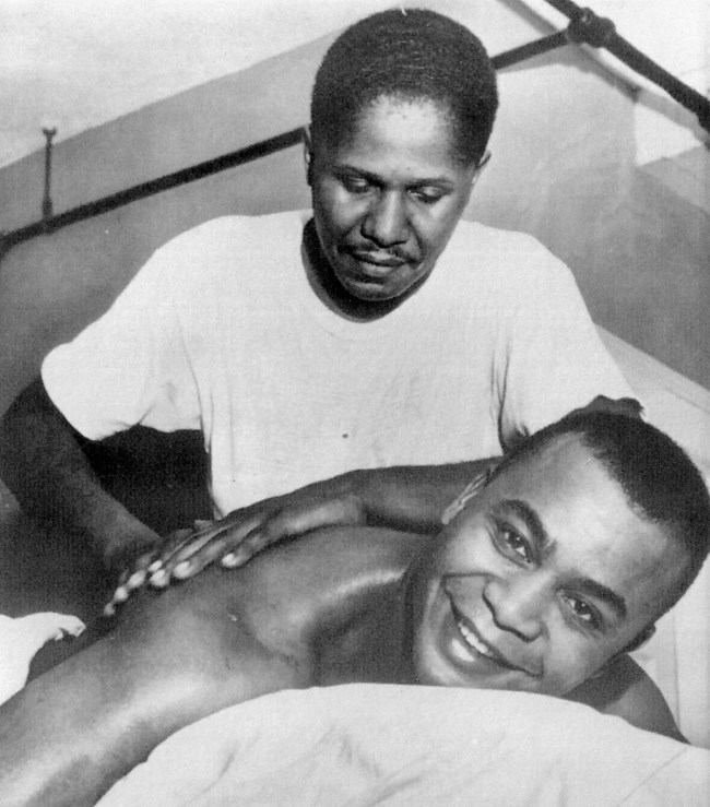 Man lays on table and smiles at camera, shirtless, while another man wearing a white shirt rubs his back