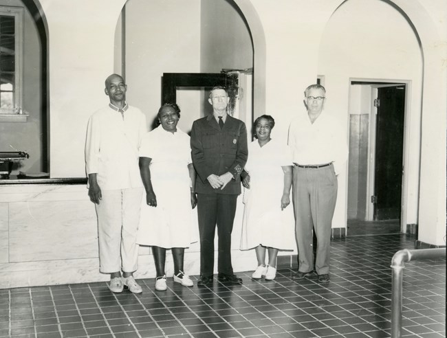 Three men and two women stand indoors. The women are wearing all-white uniforms. One man is wearing the same all white uniform as one wears a dark suit and tie and one wears a white shirt and dark pants