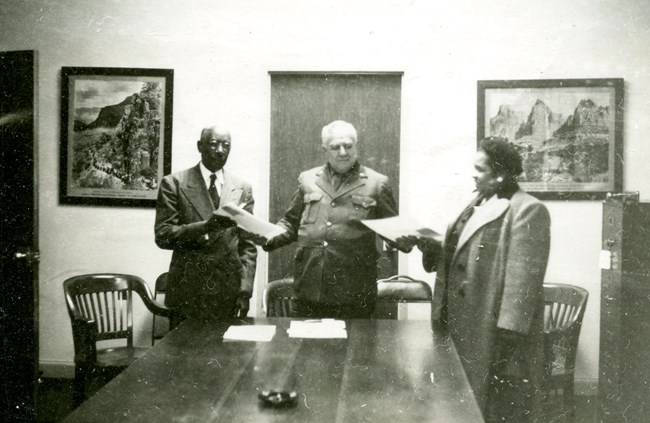 A man in a dark uniform hands paper to a man at his right and a woman at his left. All are in front of a table.