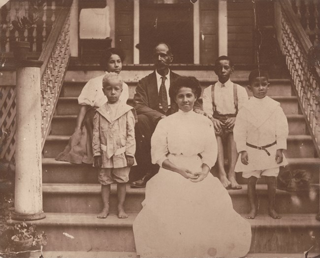 Two adults, a man and woman, and four children, three boys and one girl, sit and stand on the steps of a house