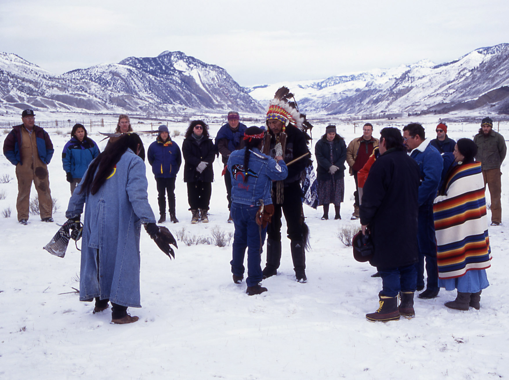 Native American people, some in traditional dress, gather with White people in a snowy mountain landscape.