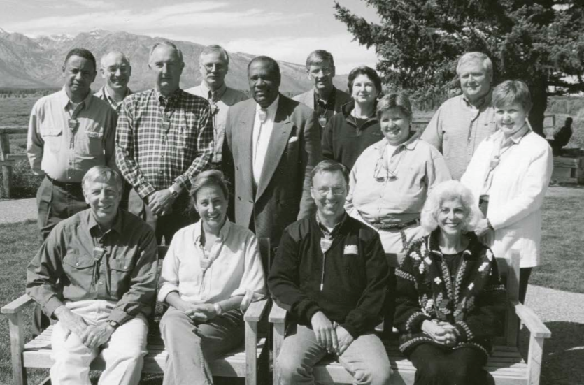 Fourteen smiling men and women pose for a portrait, mountains in background.