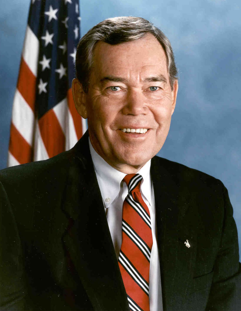 Smiling White man in black suit and red tie, American flag behind.