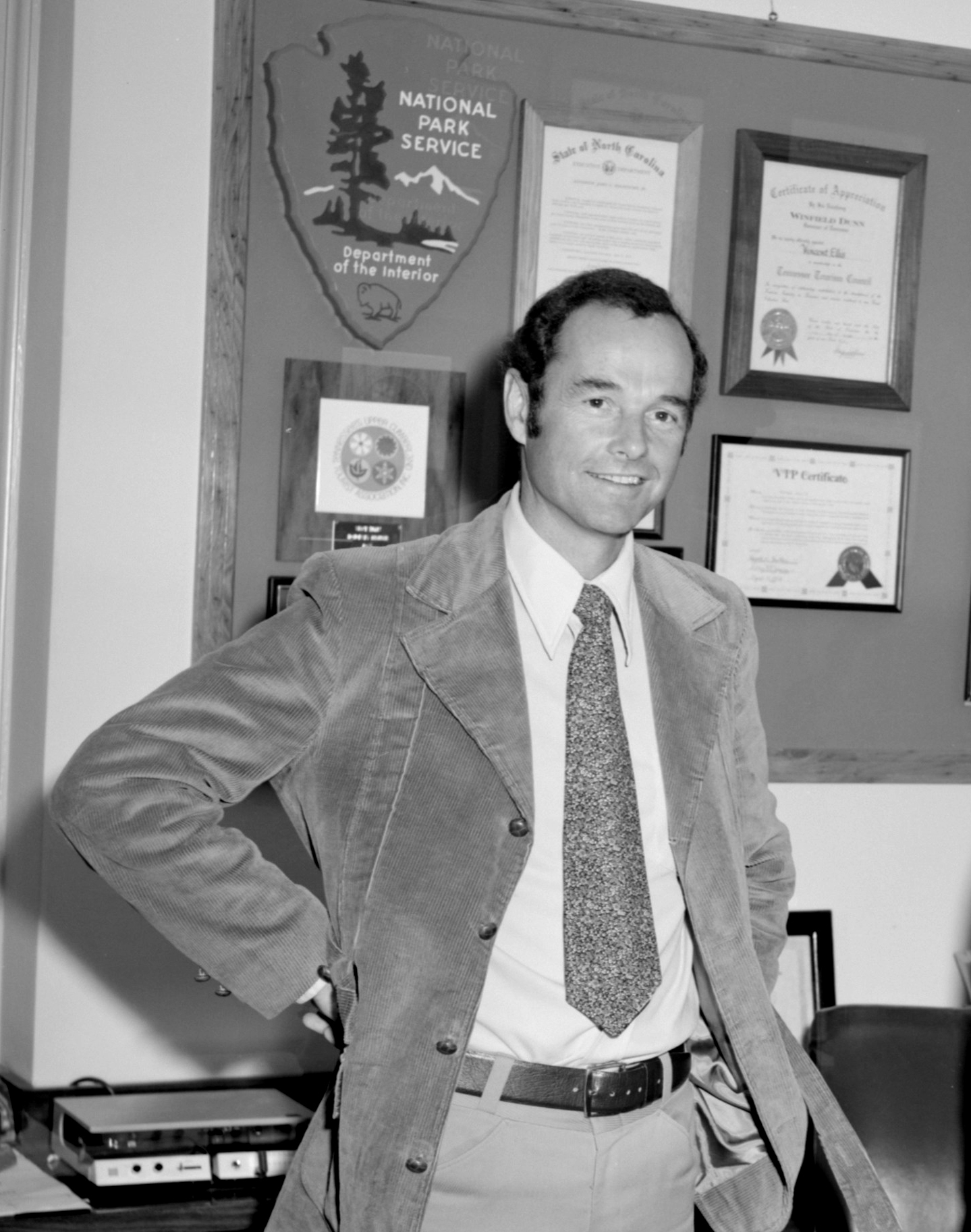 Smiling White man in corduroy jacket and wide tie stands in front of a bulletin board with awards and NPS arrowhead.