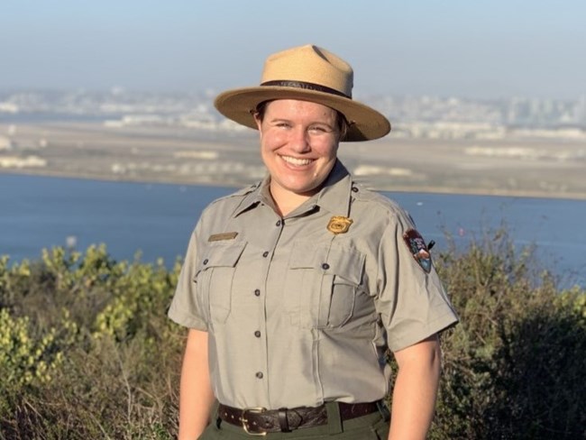 Lauren smiling outside in her National Park Service uniform and hat. San Diego Bay and the city are out of focus behind her.