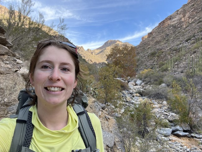 Athena Lynch, Physical Science Technician Woman wearing a daypack standing in a rocky desert wash with shrubs and saguaro cacti growing along the bottom and sides.