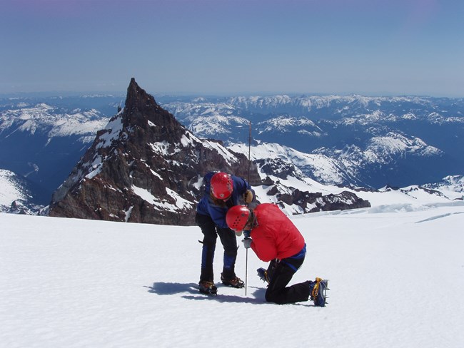 Two people in winter clothing probing a glacier to measure snow depth with snowy peaks in background