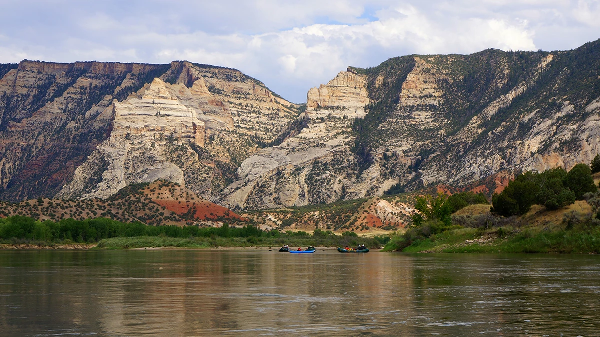 The Green River winds through Dinosaur National Monument, framed by cliffs and rugged scenery. The Green River flows below beige, brown, and red cliffs with rafters in the distance.