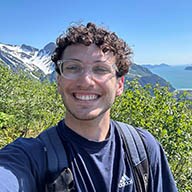 Jacob_Cohen A young white man with curly brown hair and glasses smiles on a mountain top.