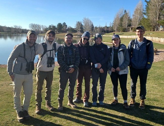 Seven smiling people all with binoculars standing in front of a pond.