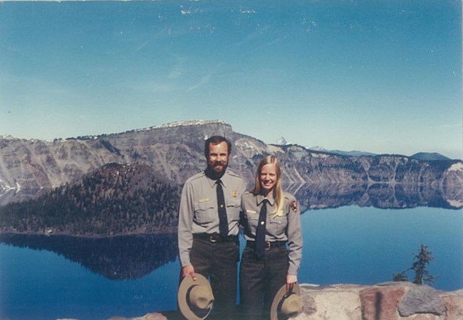 Husband and wife standing together in ranger uniforms in front of large blue lake.