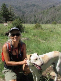 Smiling woman and dog sitting among wildflowers with forest in background.