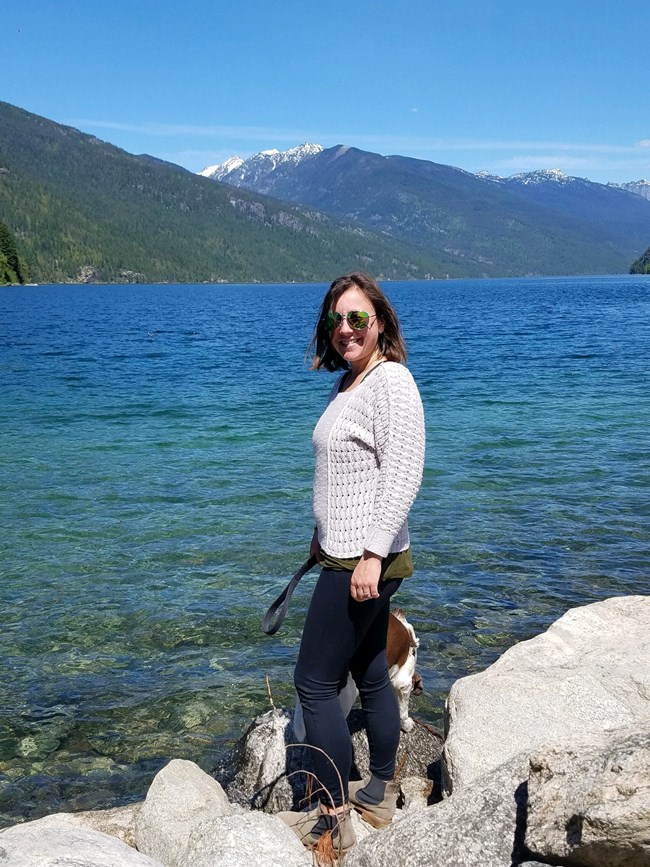 Smiling woman standing on shore of a large blue lake with her dog. Snowy mountains in background.