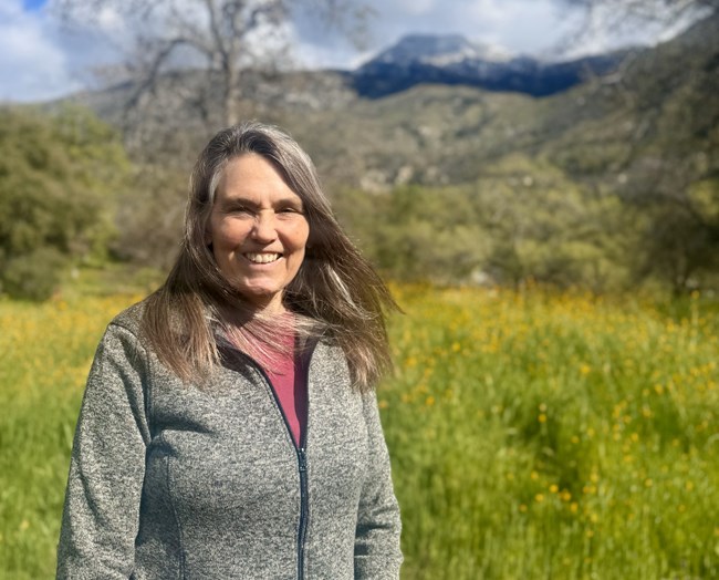Anne Pfaff, Network Program Manager A woman smiles in front of a bright green meadow with yellow flowers. A few trees just starting to leaf out and a mountain is visible in the background.