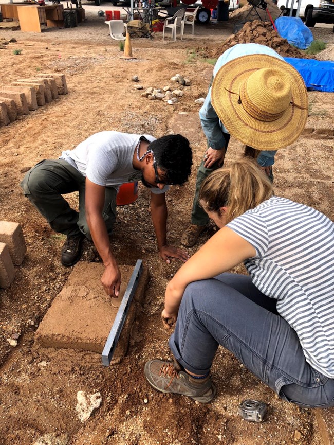 Three people stand over an adobe brick as one person uses a level to measure it