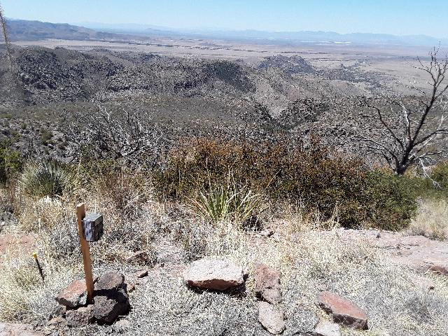 Wildlife camera on post on a brushy hillside, desert mountains in background.