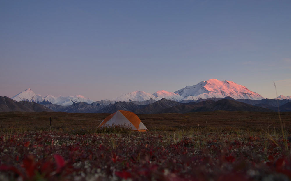 a tent set up in an open field with a view of distant mountains, including one massive mountain tinged pink by the setting sun