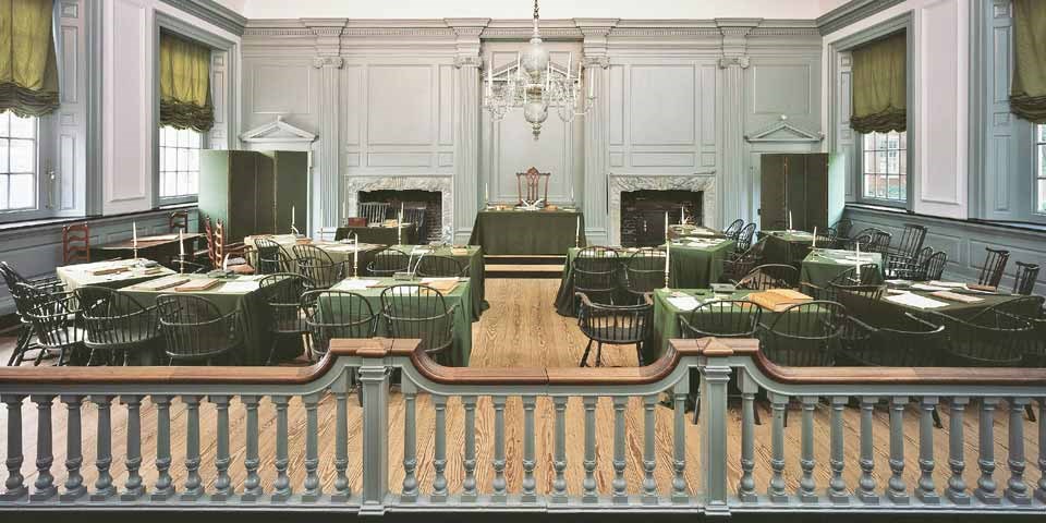 Color photo showing Windsor chairs grouped around desks covered by green cloths, all facing a large desk and chair in the front of the room.