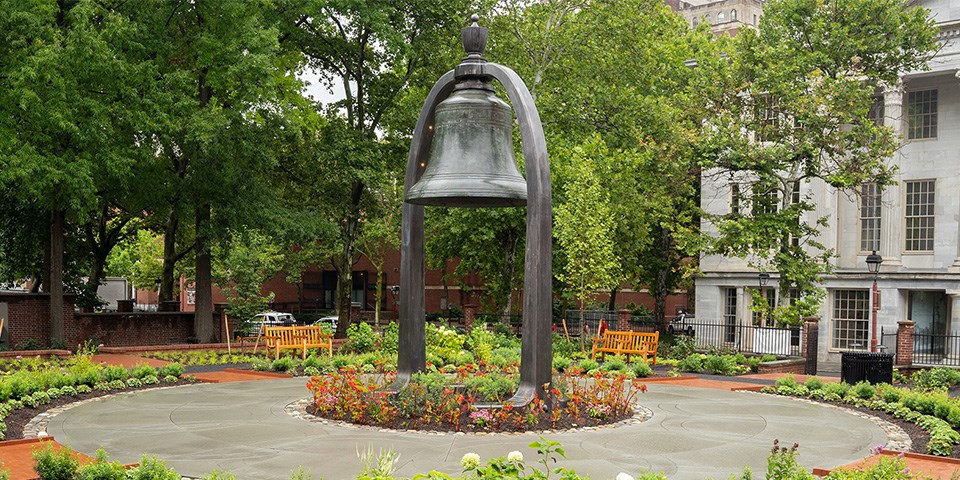 Large bell suspended by an arch surrounded by foliage.