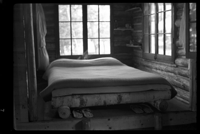 Historic photograph of the inside of the living aclove of Jun Fujita's Rainy Lake cabin. Bed supported by birch beams. Japanese sandals under bed. Log walls and large windows 