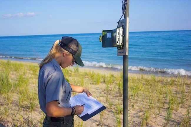 An NPS biologist stands on a sandy lakeshore next to an acoustic monitor on a pole.