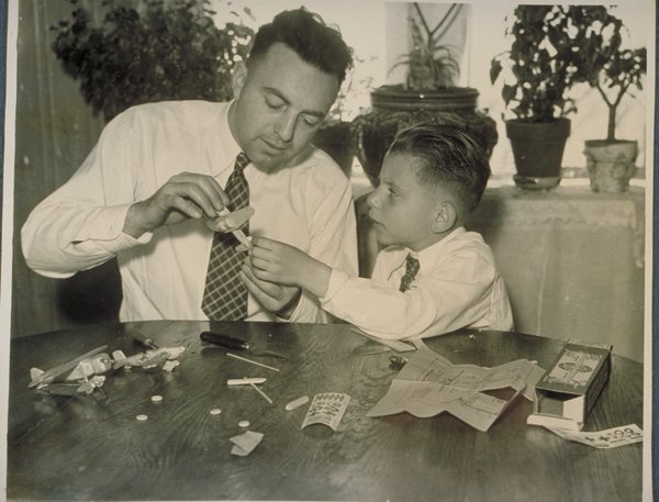 Edwin helps his son David build a model airplane while seated at a wood table inside a house.