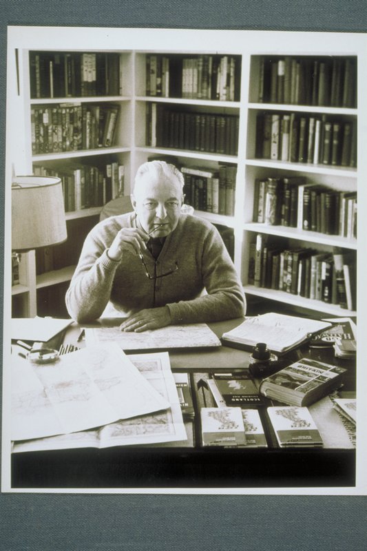 A seventy-year-old man sits at a desk with papers and books; book shelves are behind him.